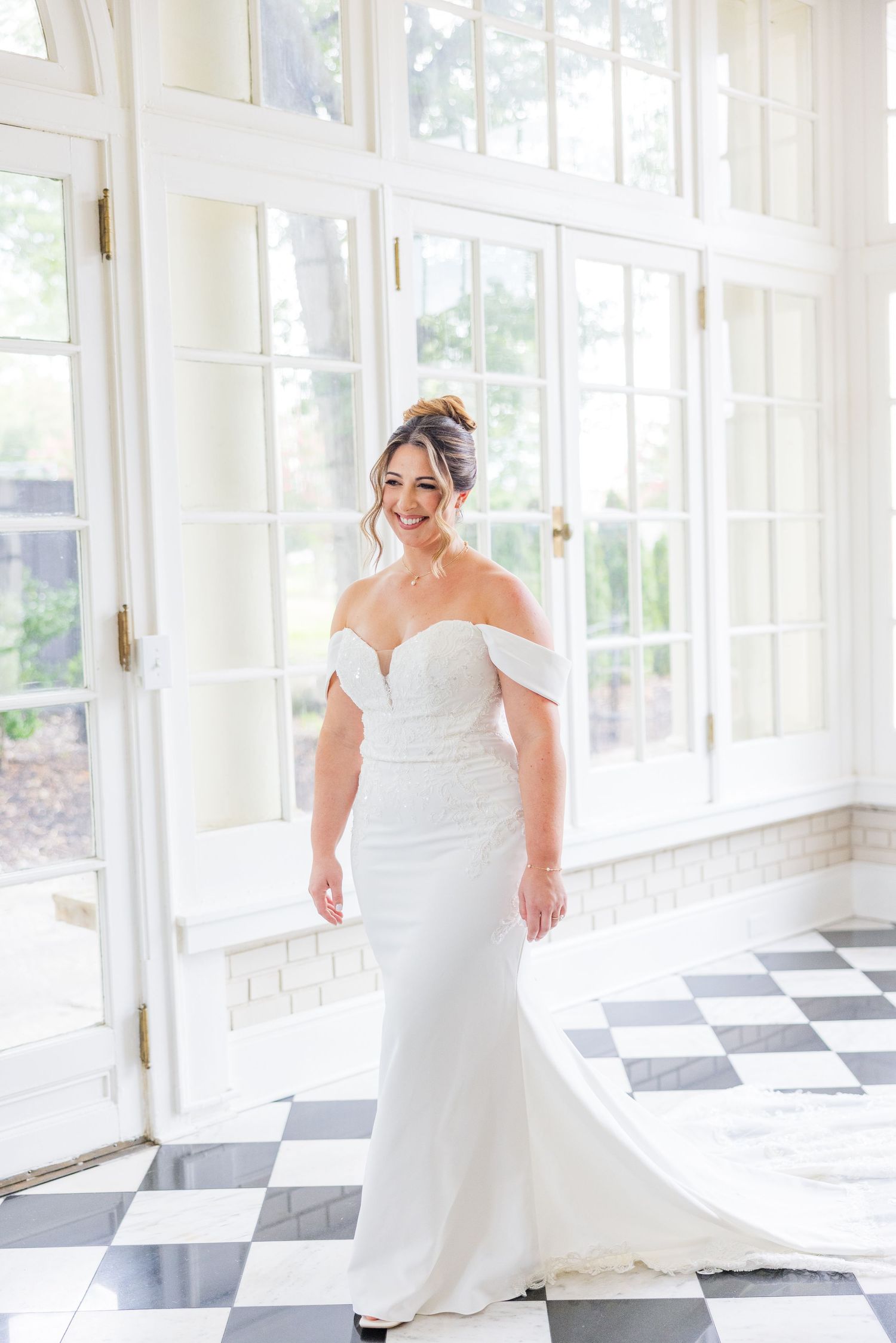 A bride in a flowing white dress stands on a black and white checkered floor in a sunlit room.