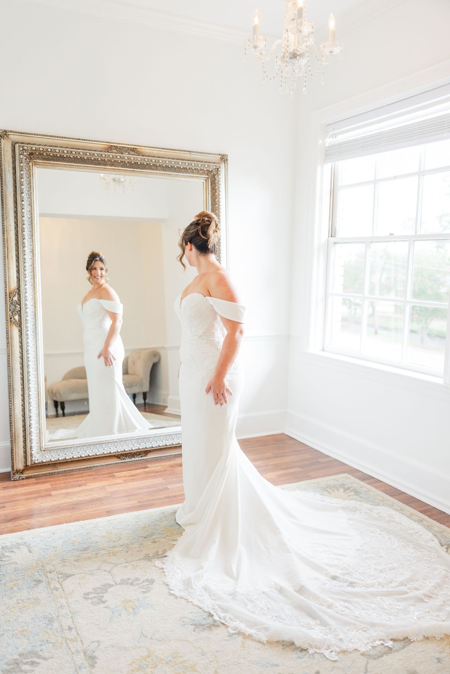 Bride in flowing white gown twirls and poses in front of large ornate mirror in bright room.
