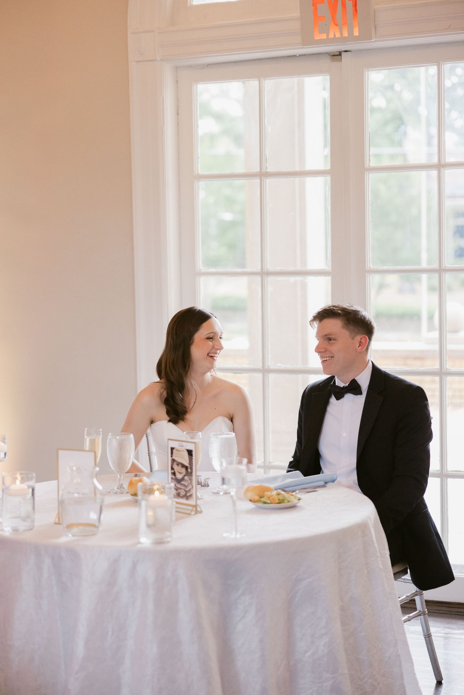 A wedding reception setup with white tablecloths and elegant place settings in a bright, window-filled room.