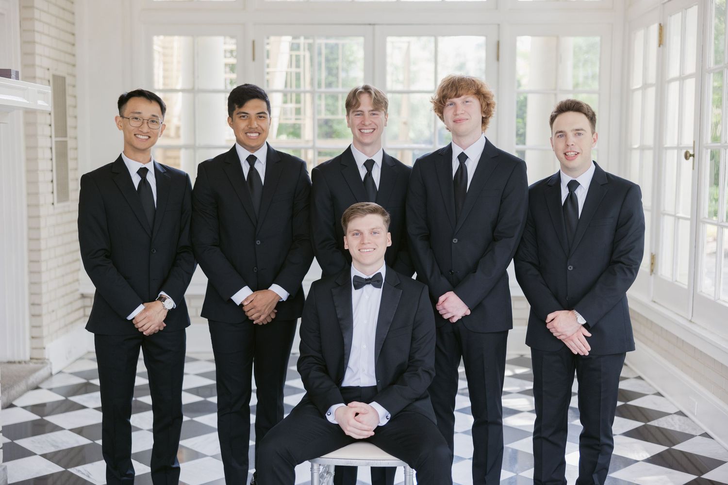 A group of groomsmen in black suits pose together in an elegant hallway with checkered floors and white columns.