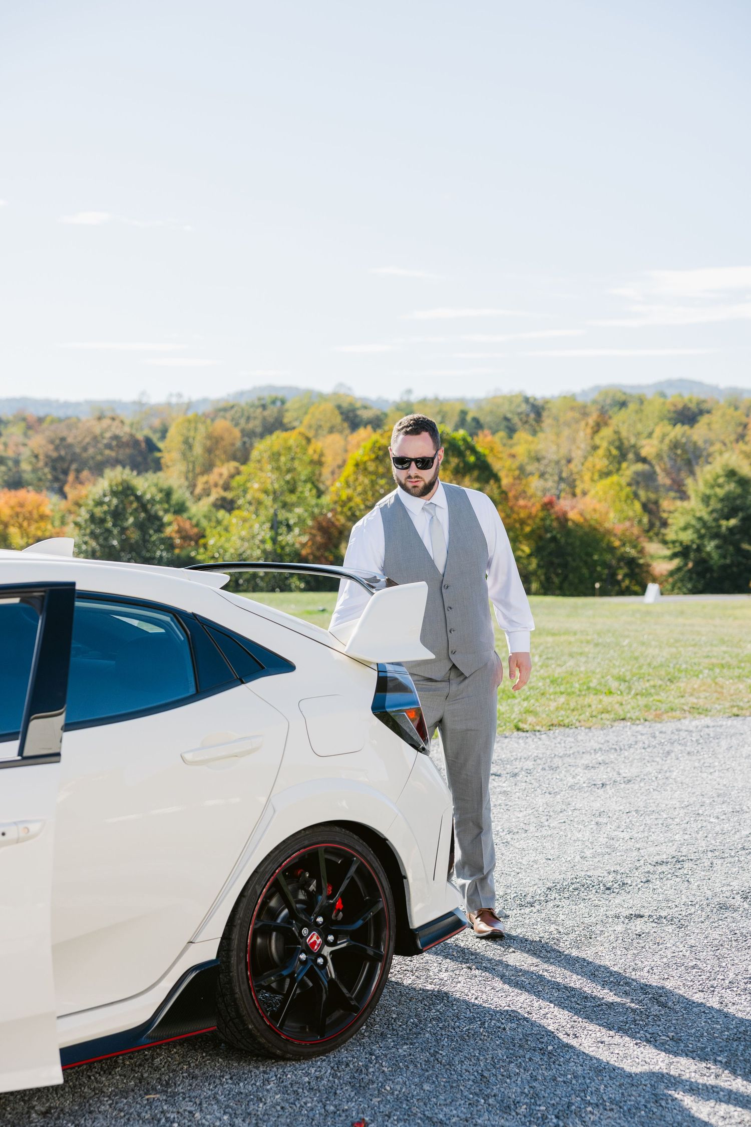 White sports car parked on gravel with wedding party members in formal attire posing nearby.