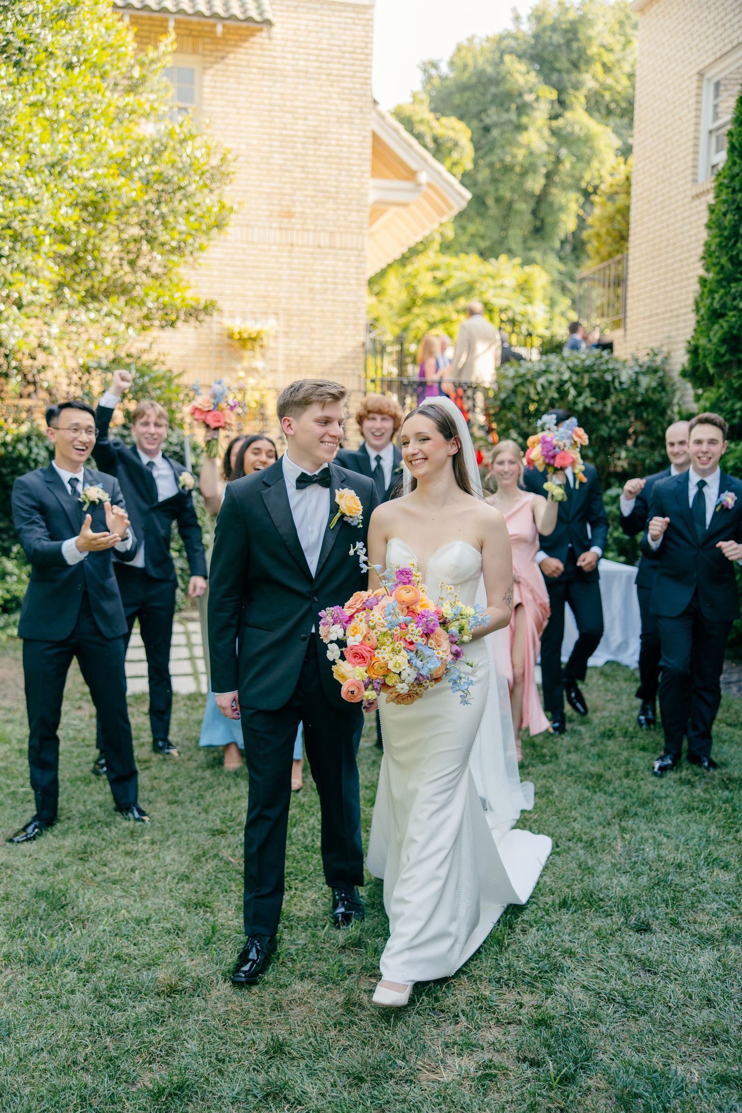 Wedding party celebrates outdoors on lawn with guests in formal attire and colorful bouquets.