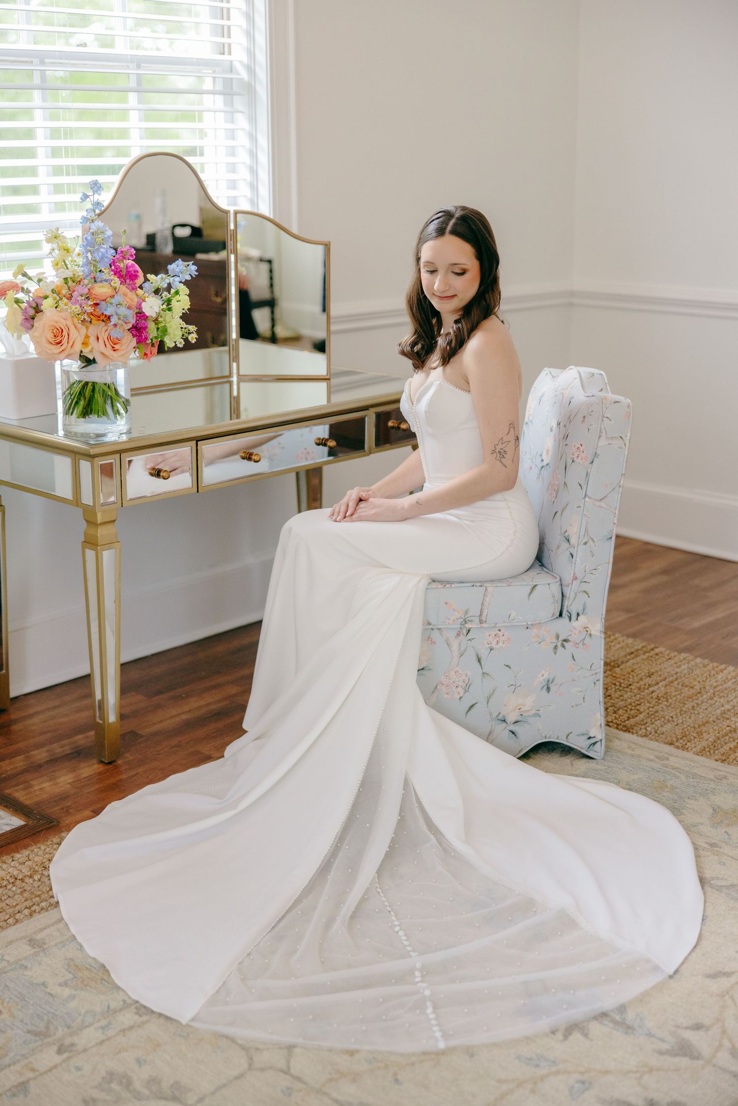 A white wedding dress flows elegantly around a floral upholstered chair in a bright room with hardwood floors.