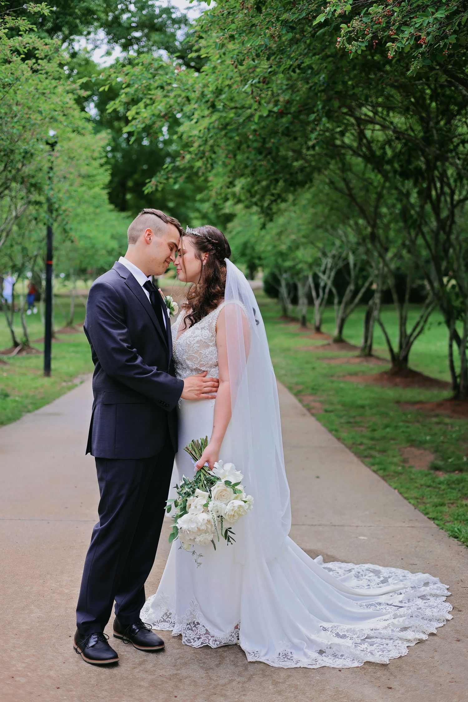 A romantic wedding moment captured on a tree-lined path with the bride's lace dress trailing behind.