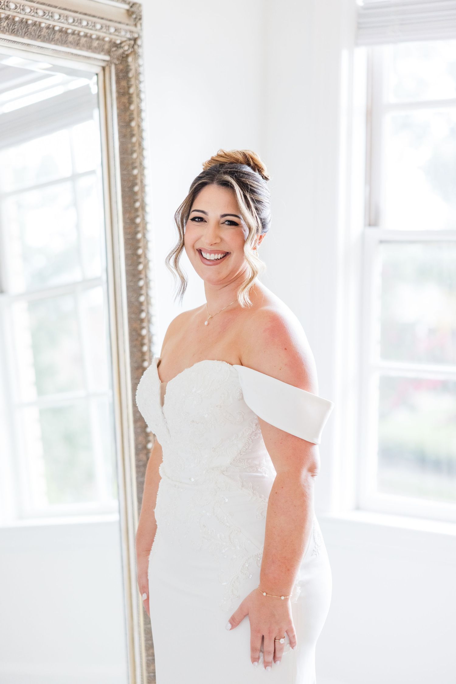 A bride in an elegant off-shoulder white wedding gown poses in front of an ornate mirror in a bright room.