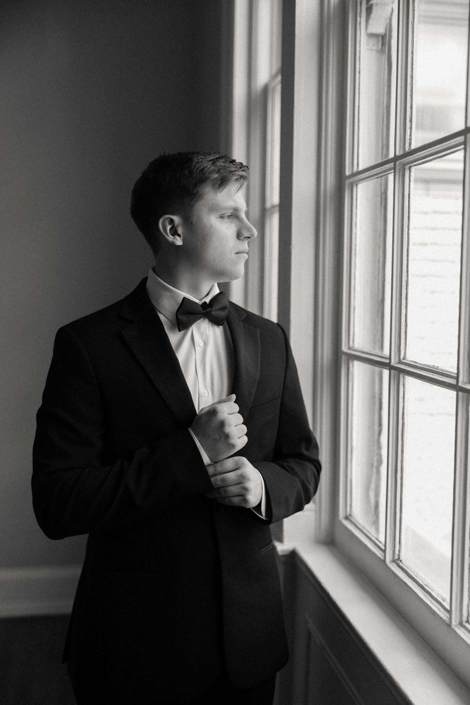 Classic black and white portrait of groom in tuxedo gazing out window.