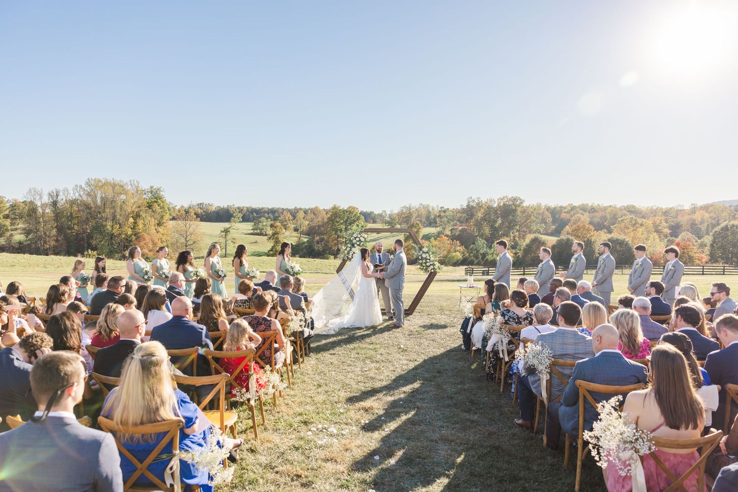 An outdoor wedding ceremony takes place on a grassy hillside with guests seated in rows on a sunny autumn day.