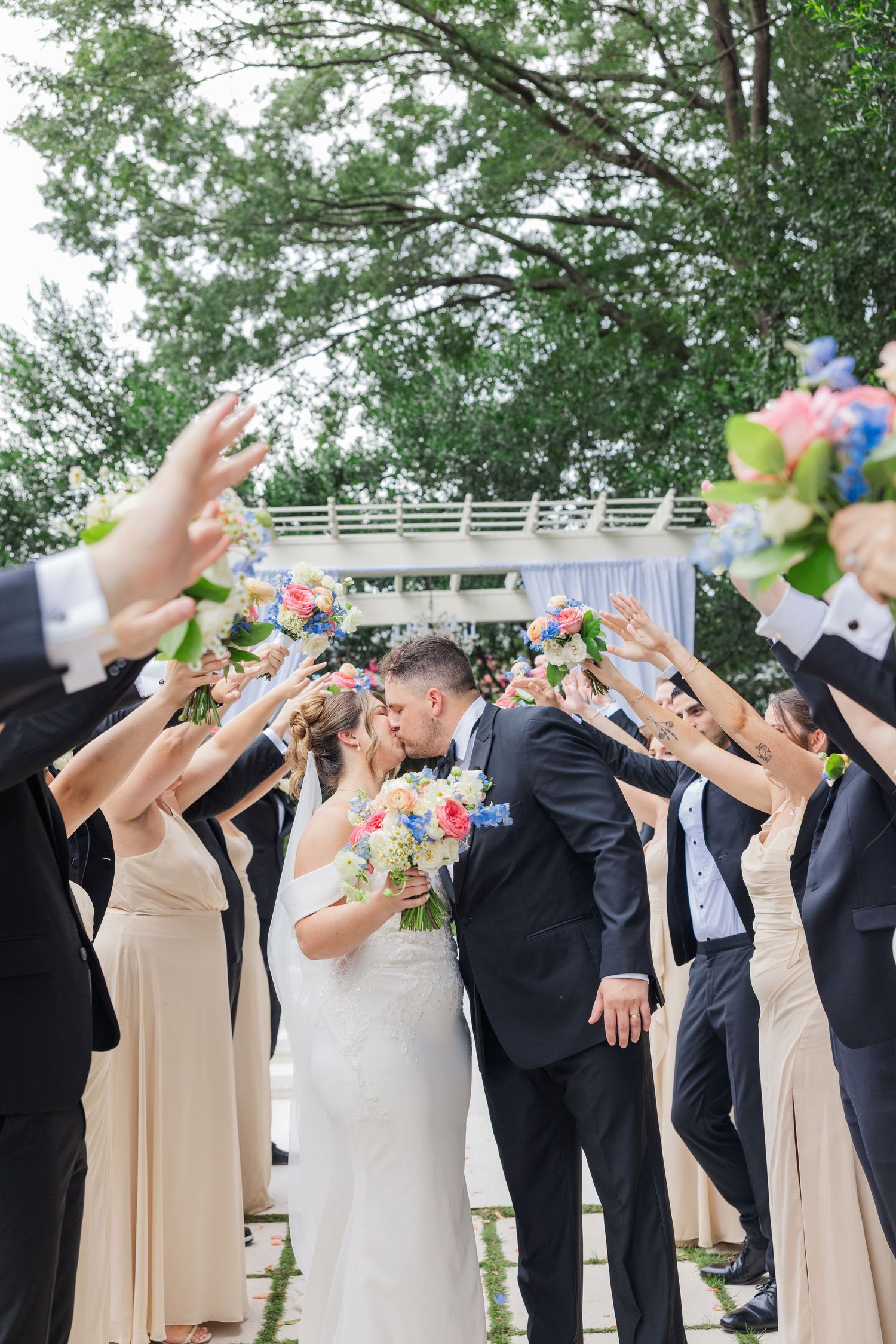 Wedding party forms a line outdoors with raised arms in celebration under green trees.