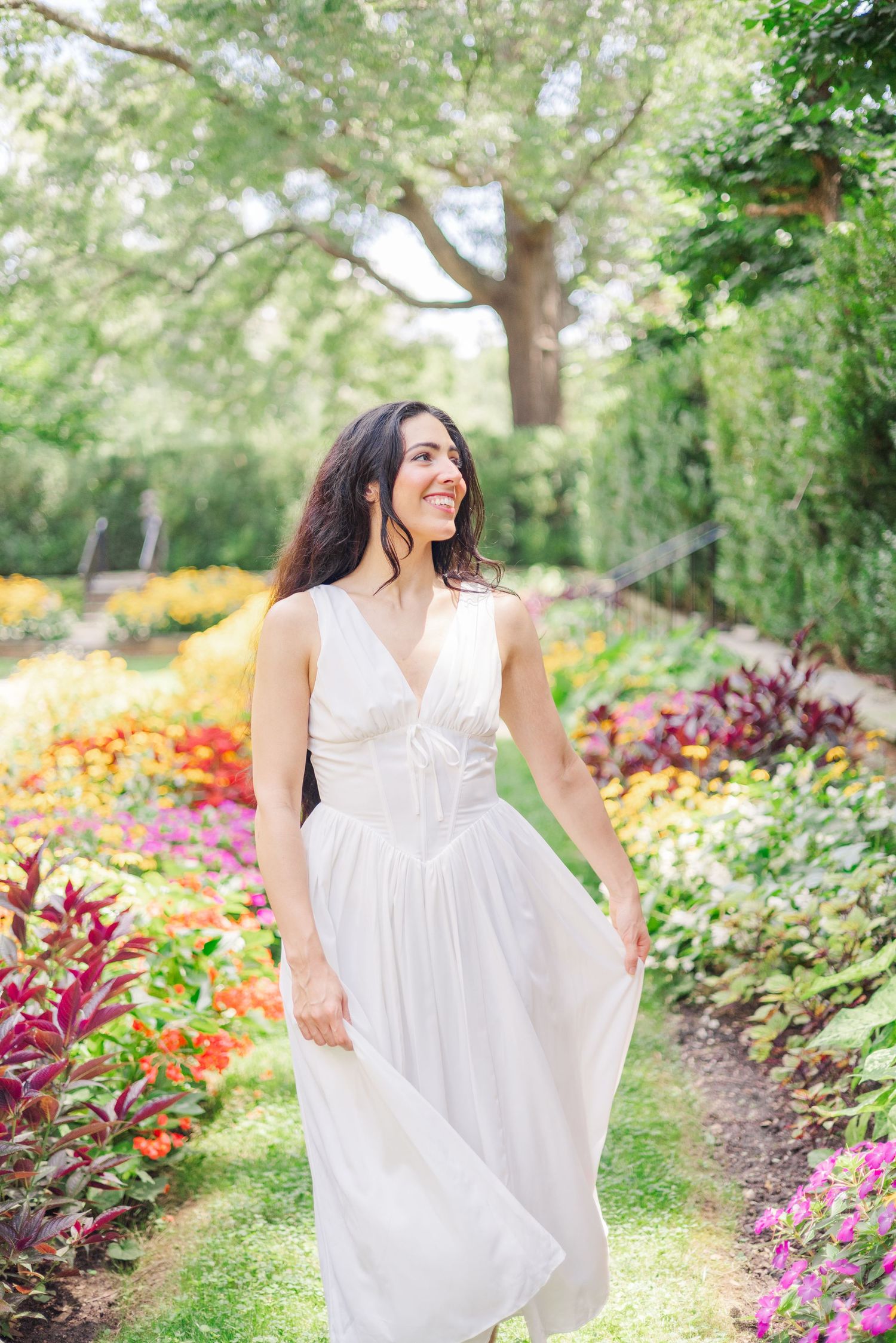 A person in a white sundress twirls and dances along a garden path bordered by yellow and red flowers.