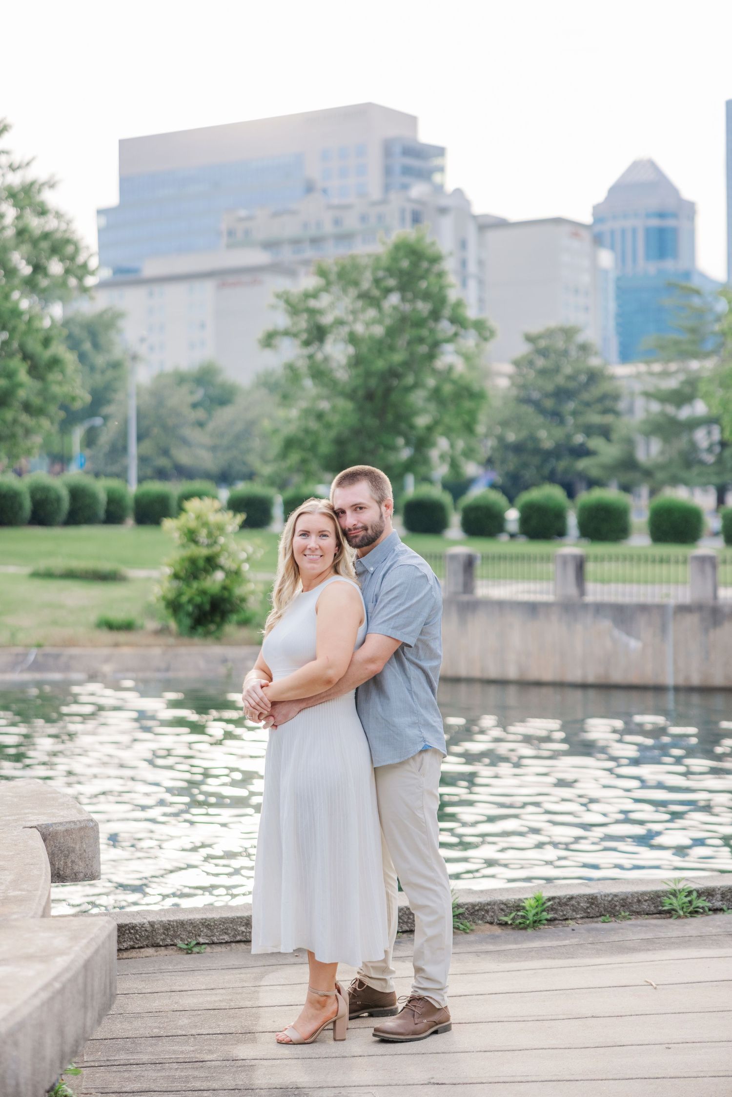Romantic couple embraces by a scenic pond with urban skyline and water lilies in view.