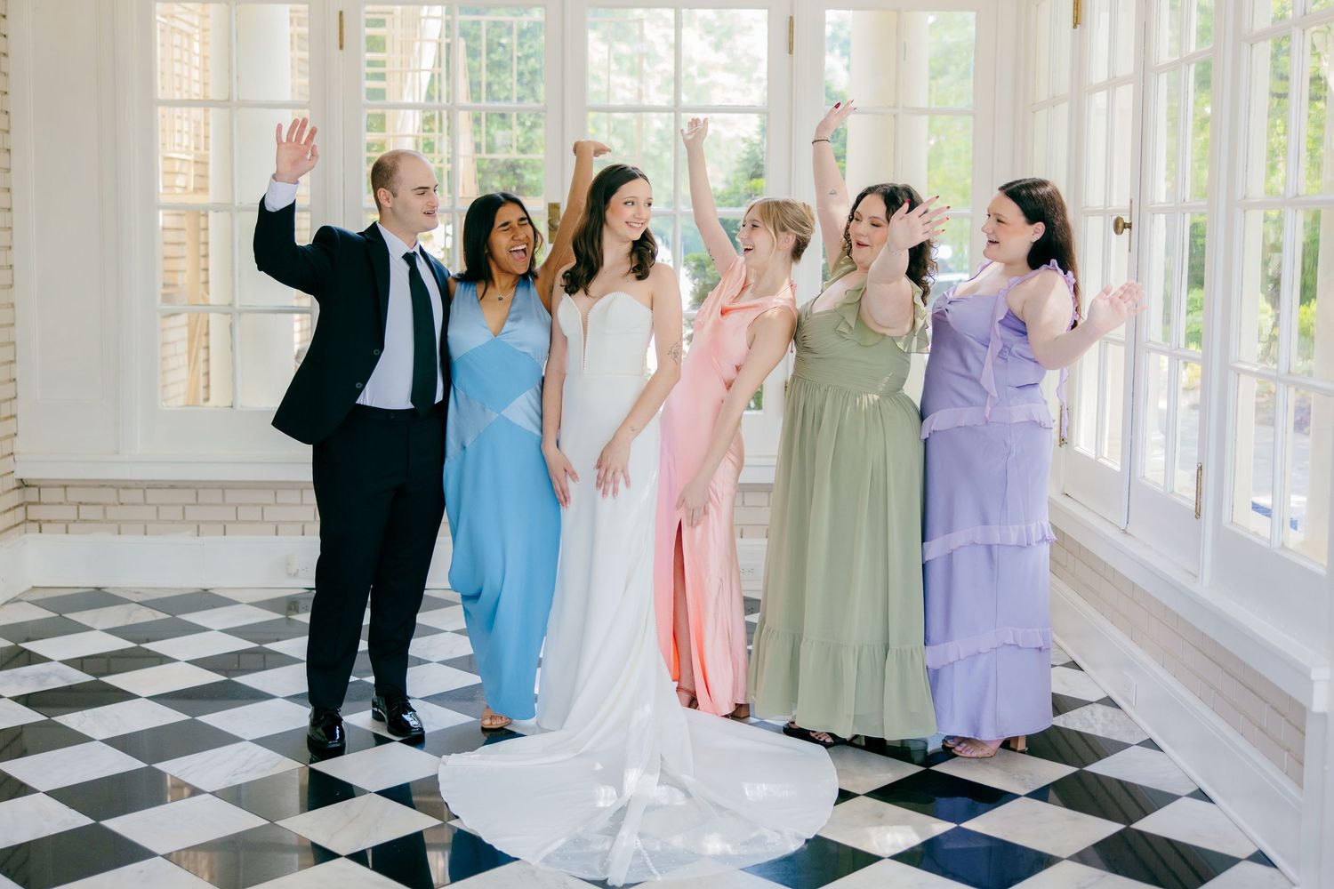 Wedding party in pastel dresses and dark suits posing together in a bright sunlit venue with checkered floors.