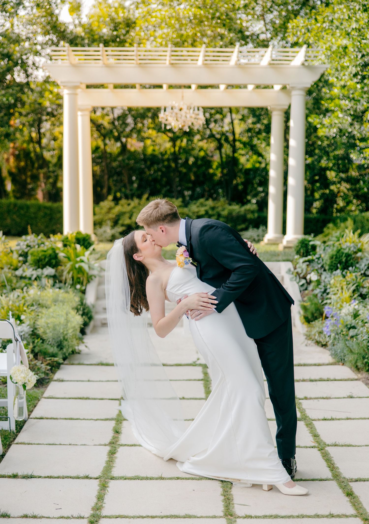 Romantic wedding couple dancing and dipping under white garden pergola surrounded by flowers.