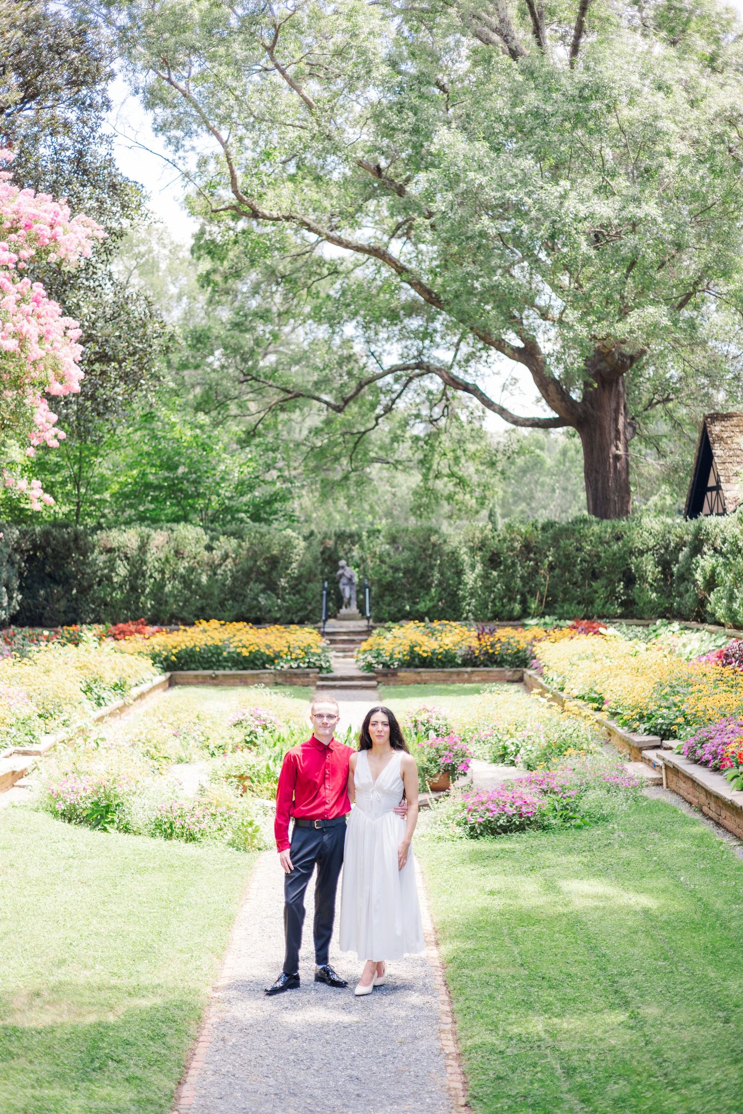 A couple strolls along a garden path surrounded by trees and flowering plants.