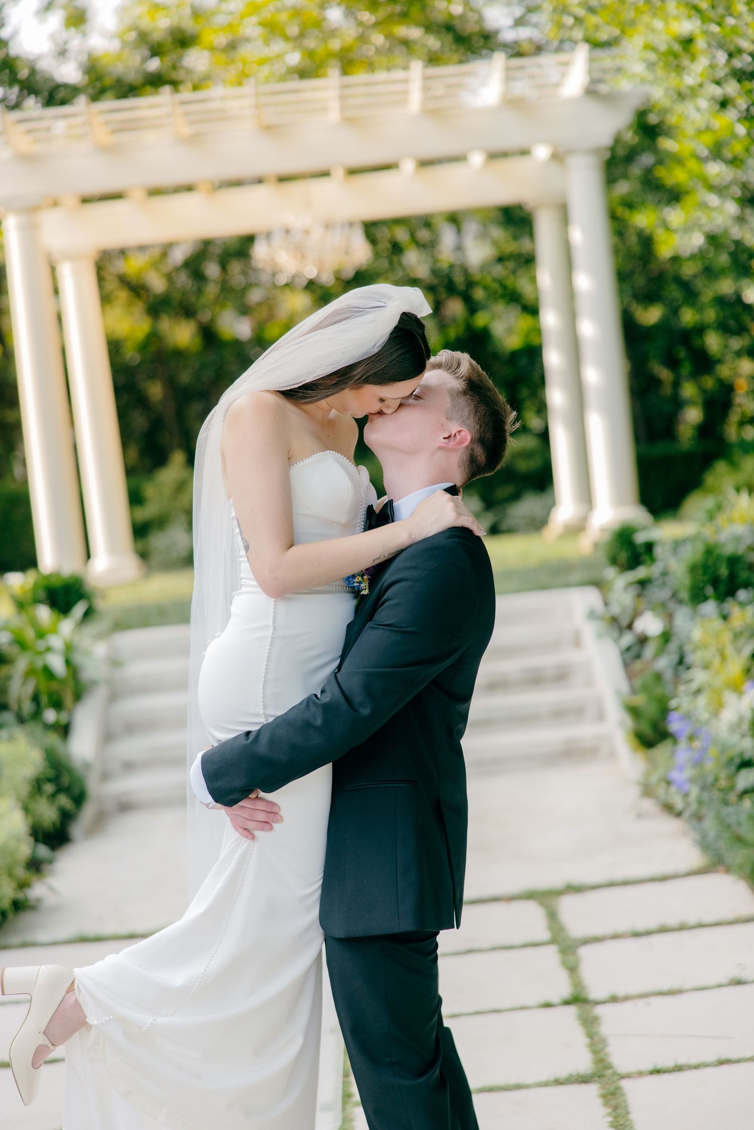 Romantic moment captured in a garden setting with white pergola and stone pathways.