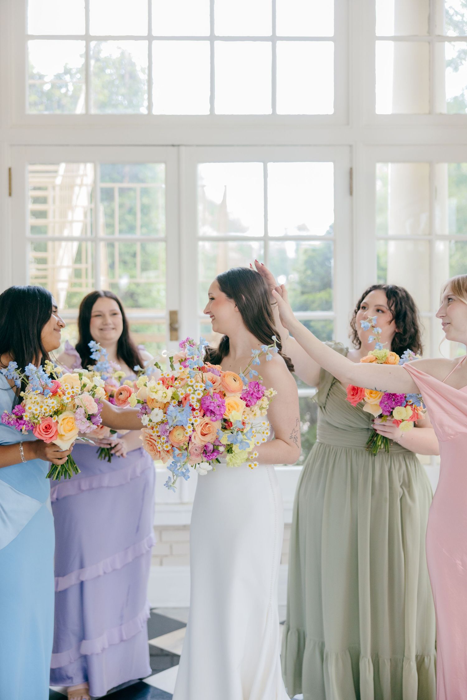 Bridesmaids in pastel dresses hold vibrant floral bouquets while posing in a sun-filled room.
