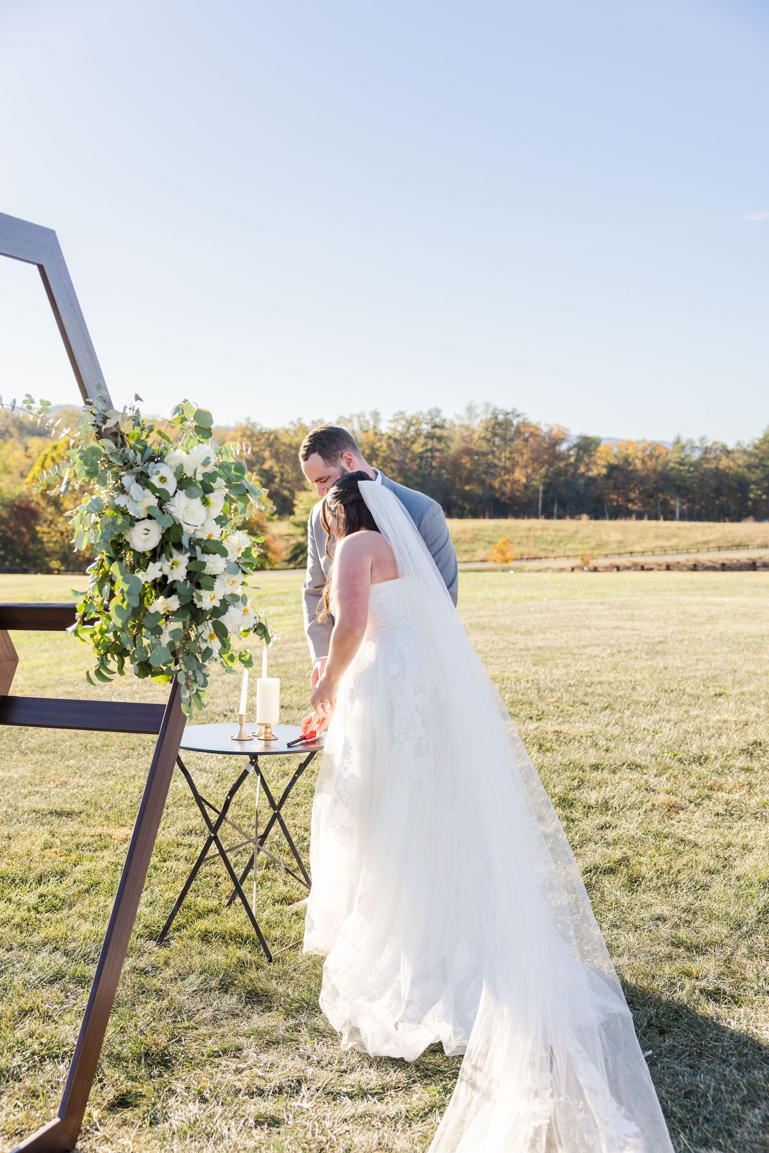 Outdoor wedding ceremony space with wooden geometric arch and white floral arrangements in a sunny field.