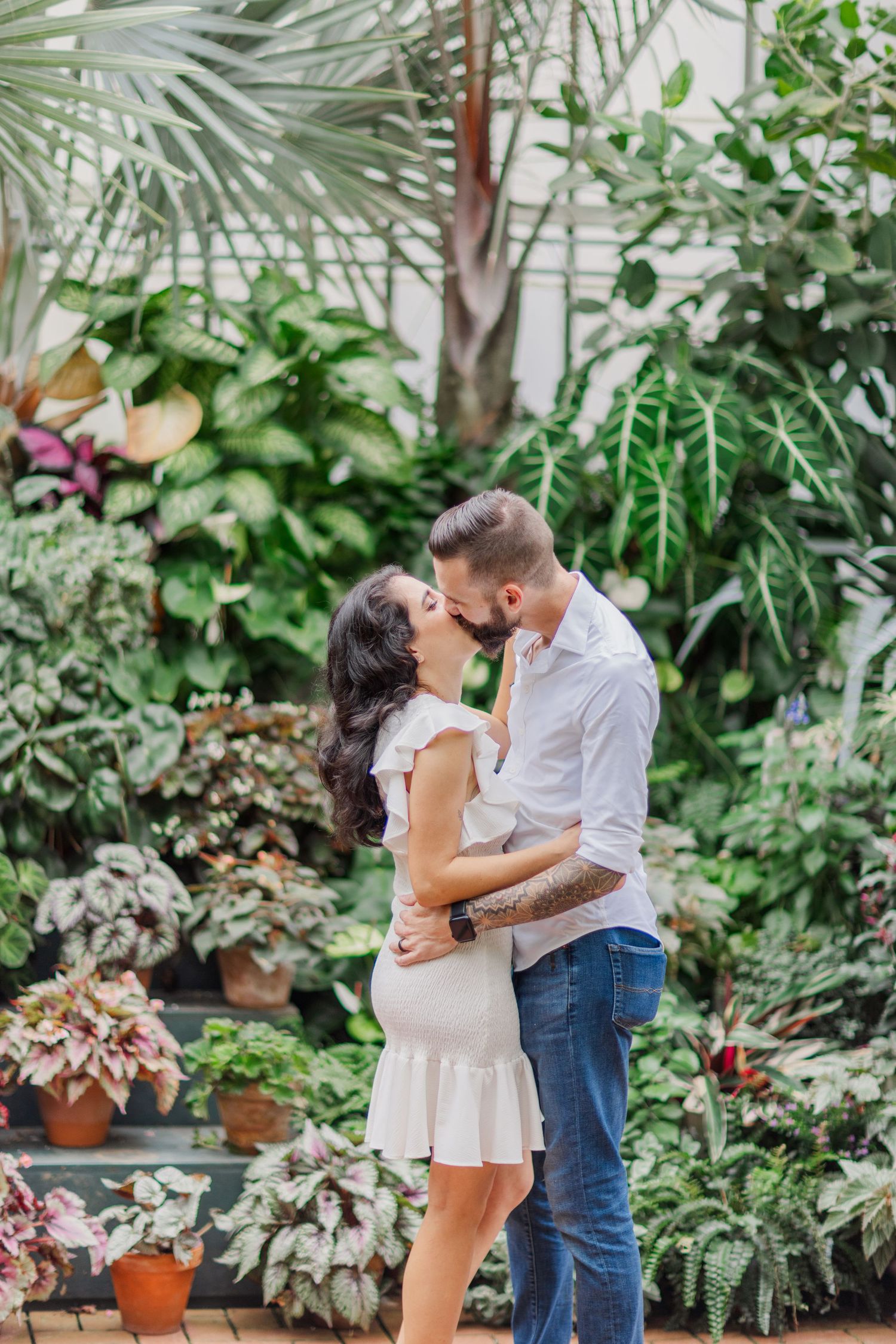 A romantic moment captured in a greenhouse setting with potted plants and tropical foliage.