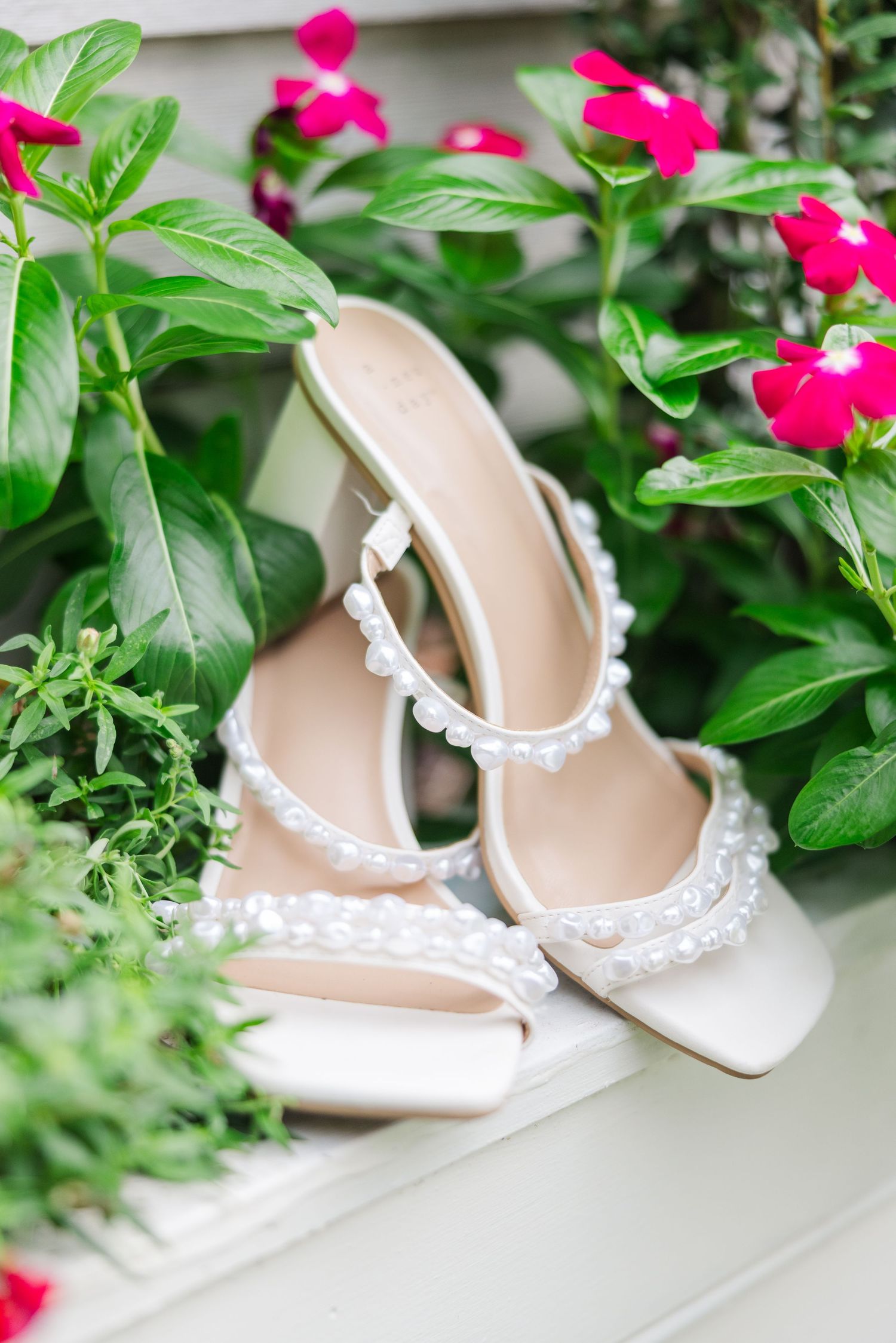 Delicate white bridal shoes with pearl straps displayed among bright pink flowers and greenery.