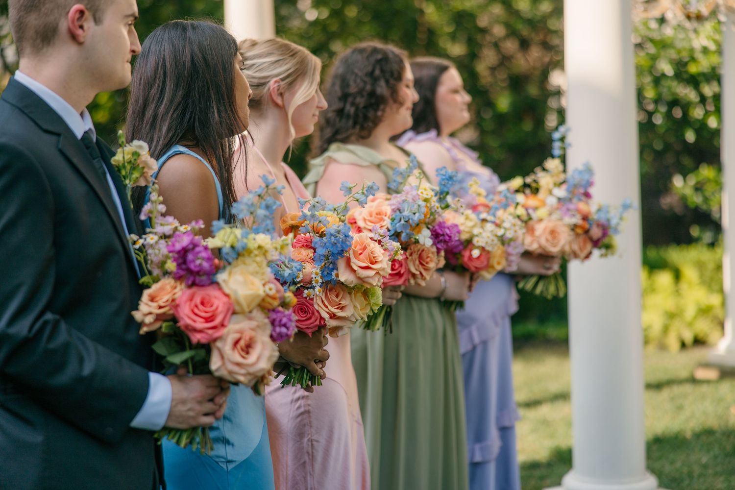 Wedding party holding colorful bouquets while standing under white columns outdoors.