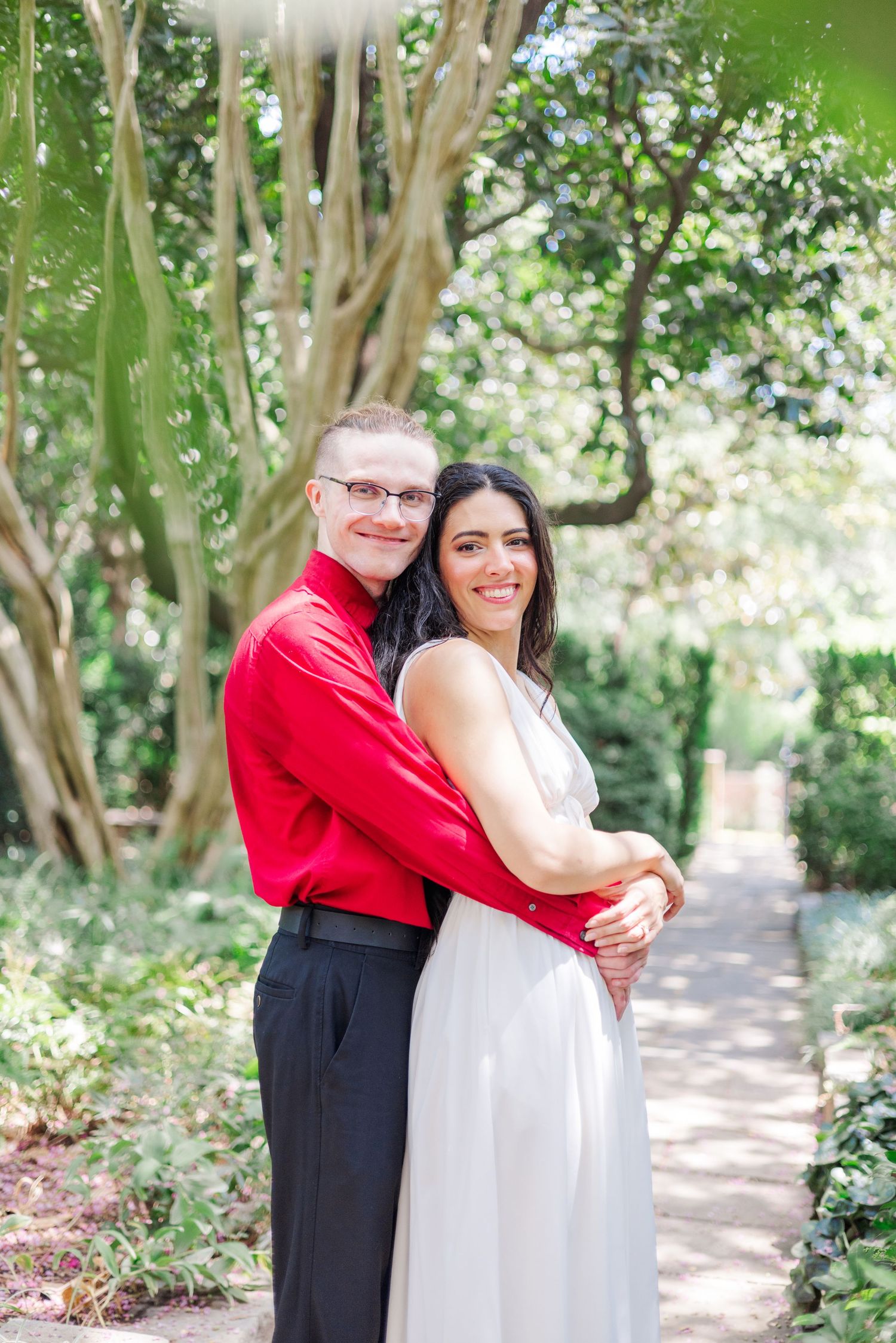 A tender embrace captured on a sunlit garden path surrounded by lush trees and foliage.