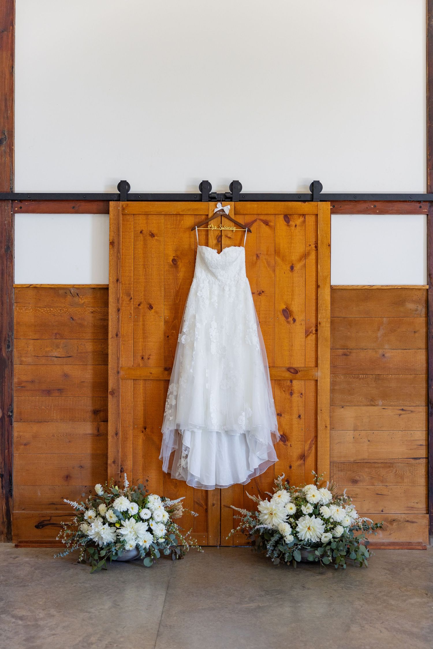 White wedding dress displayed against rustic wooden barn doors with elegant floral arrangements at the base.
