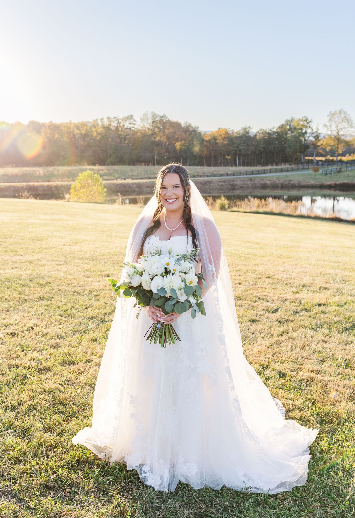 A bride in a flowing white dress poses with her bouquet in a sunlit field during golden hour.