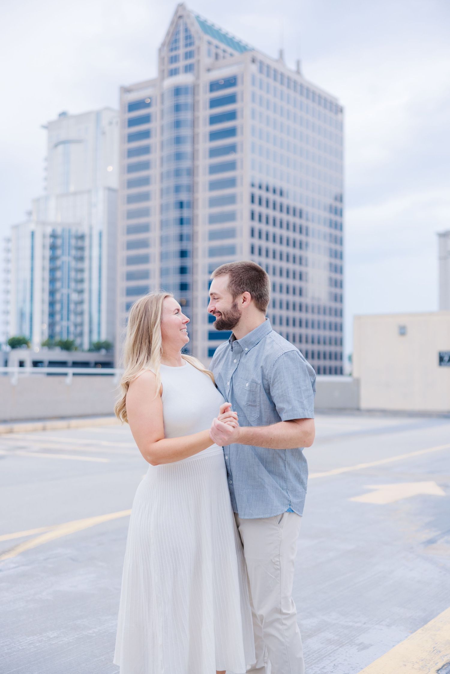 A couple in light summer attire poses playfully against an urban backdrop of modern office buildings.