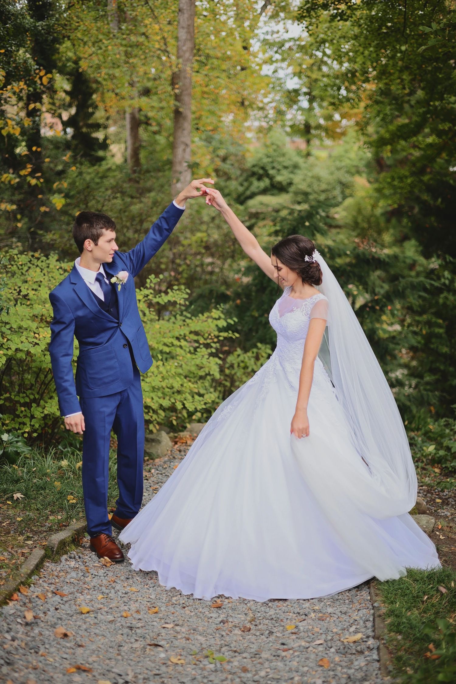 A couple in wedding attire dance together on a gravel path surrounded by autumn trees and greenery.