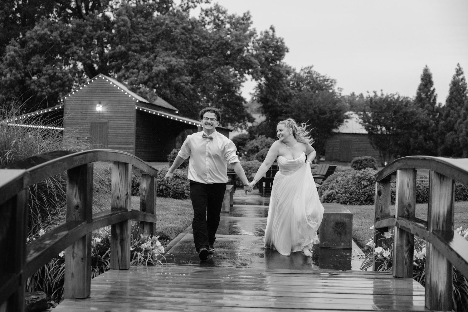 Black and white photo of a bride and groom holding hands, walking on a wooden bridge at an outdoor wedding venue.
