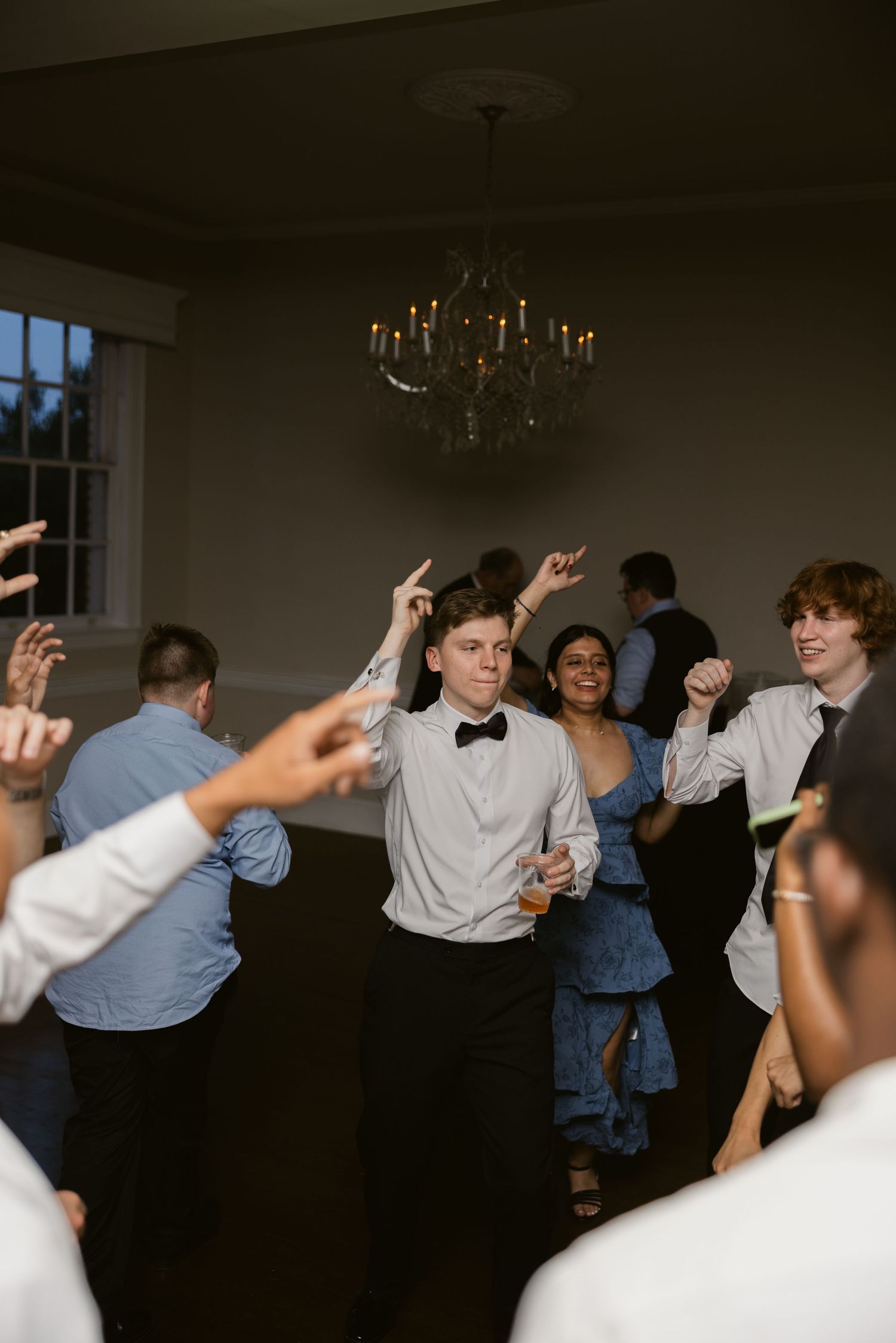 Wedding reception guests dancing with raised arms in a dimly lit room.