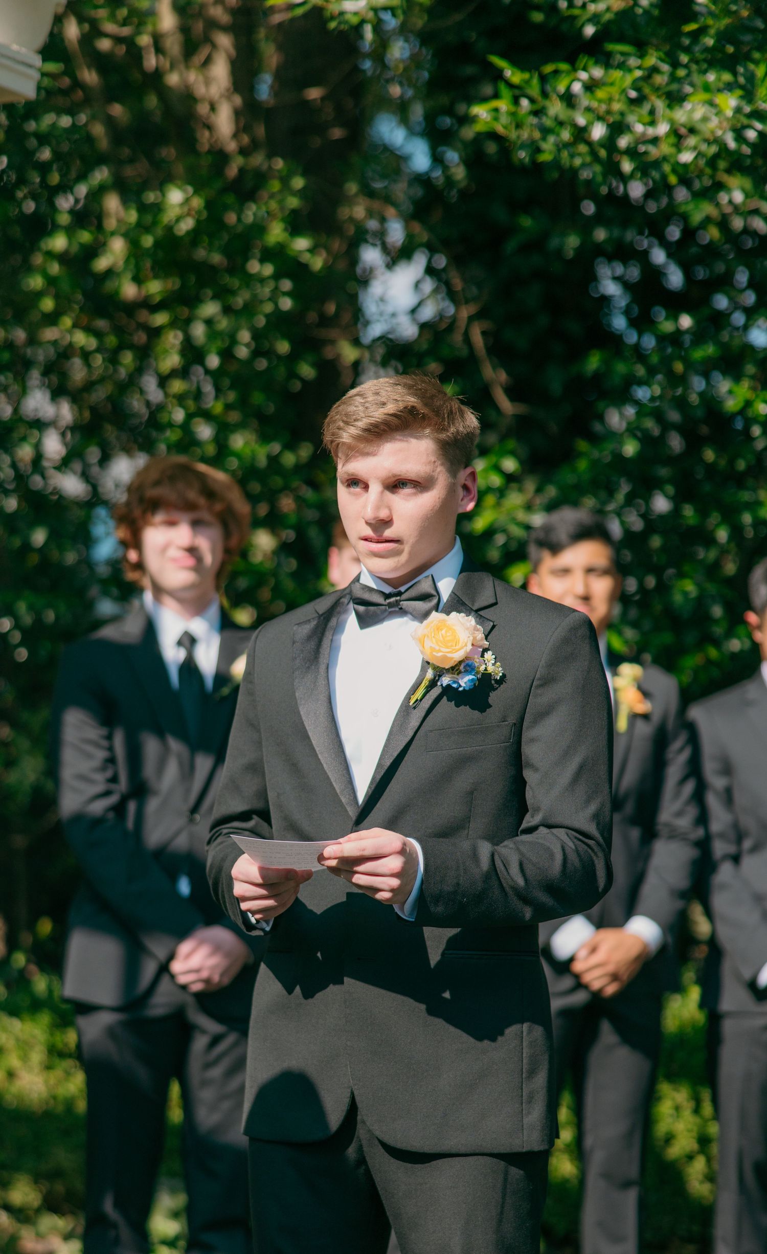 Outdoor wedding scene with groomsmen in dark suits standing near trees.