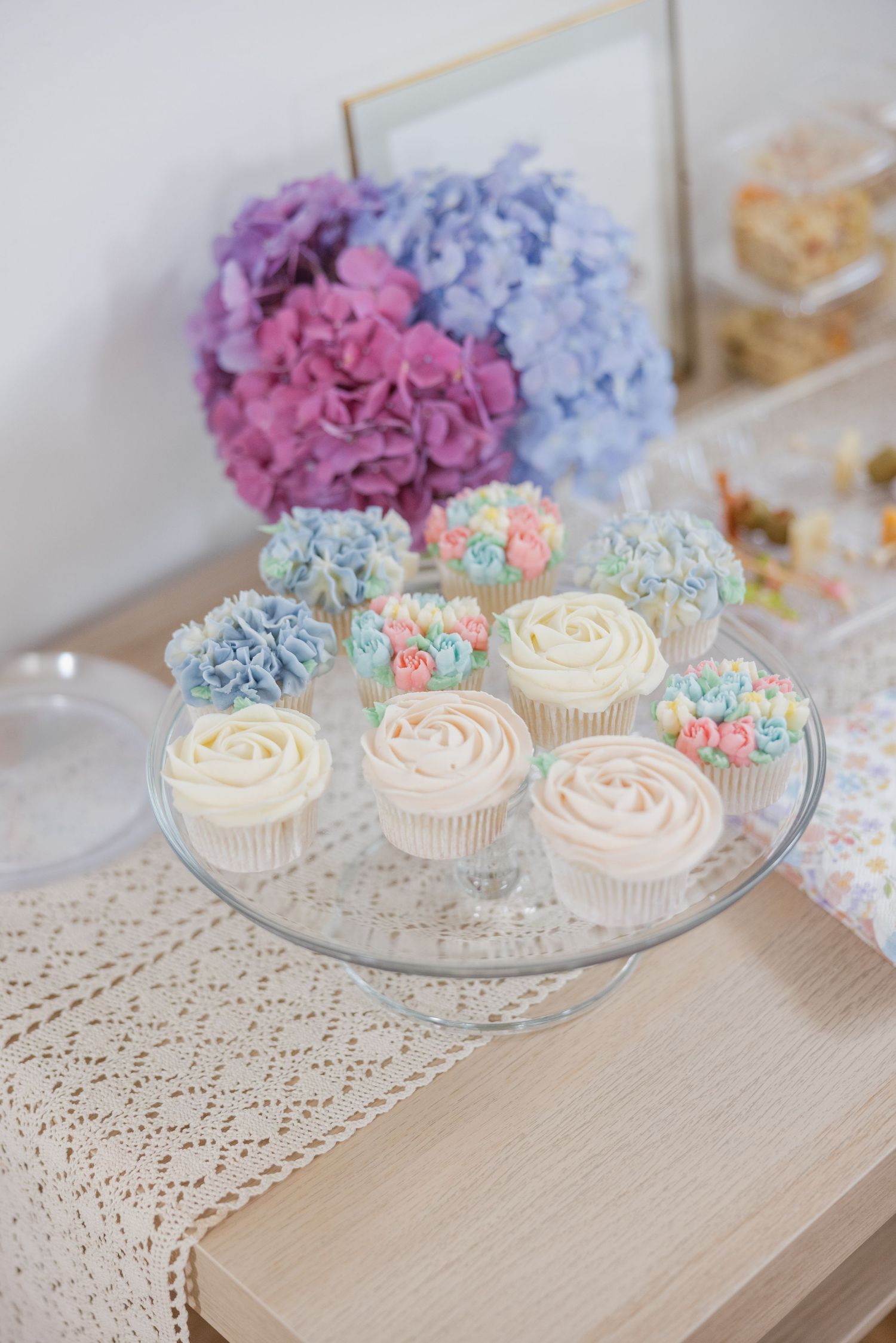 Delicate cupcakes with pastel frosting displayed on glass stands alongside purple and blue hydrangeas.