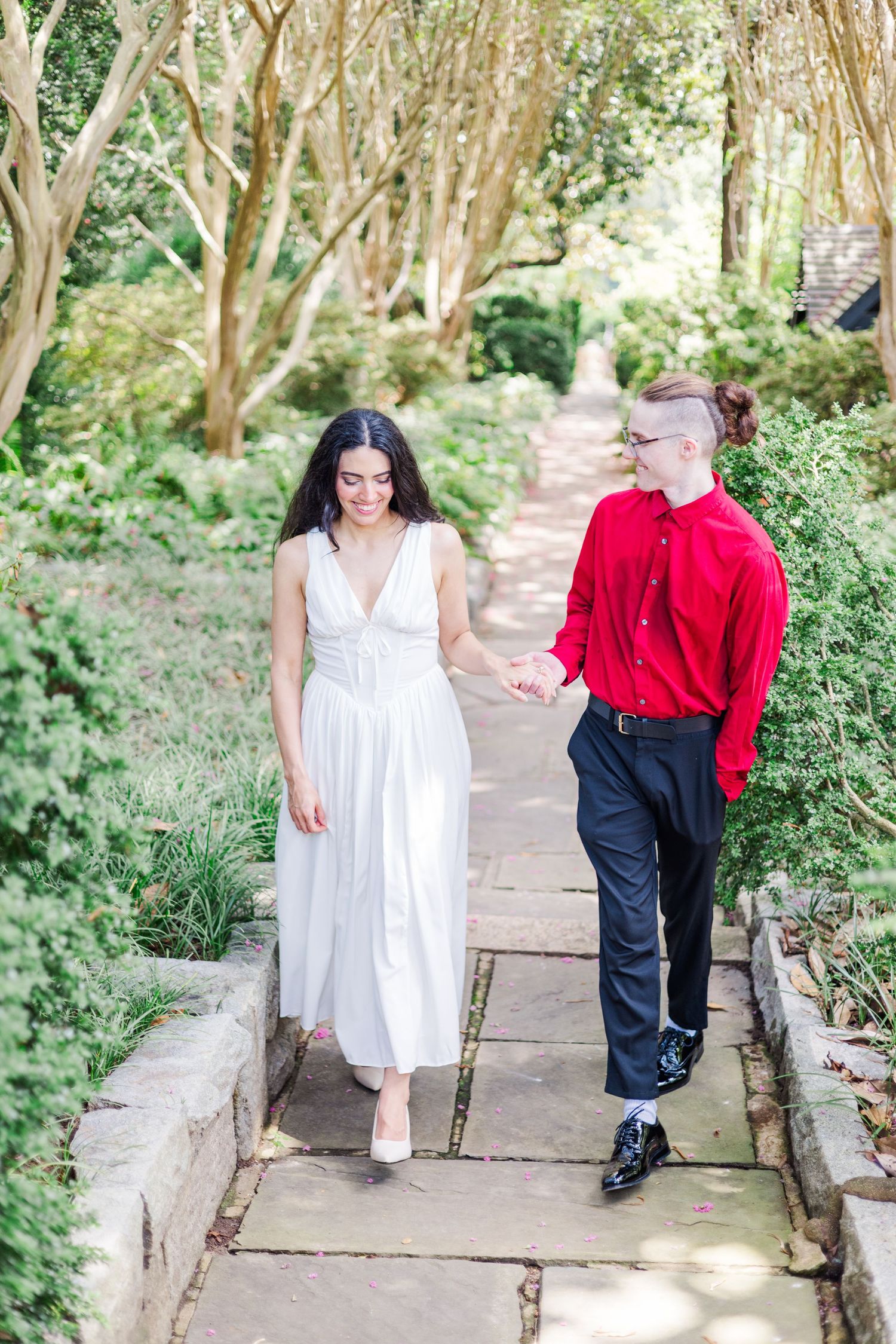 A couple walks hand in hand along a stone path surrounded by trees and greenery.