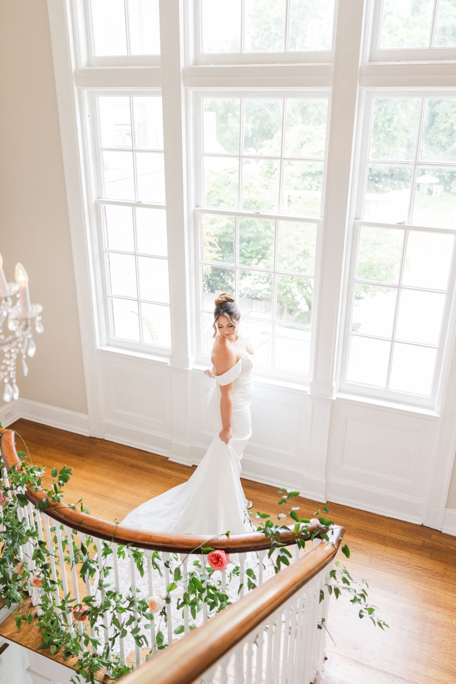 Elegant white bridal gown displayed in sunlit room with floral garland decorations along wooden railing.