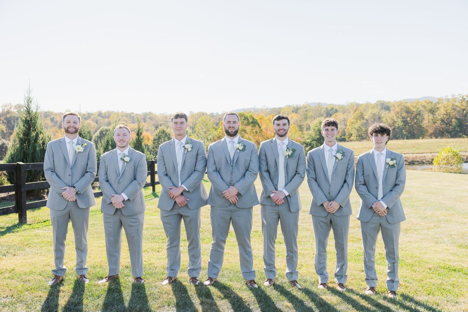 A wedding party of groomsmen wearing matching light gray suits stand in a line outdoors on a sunny day.