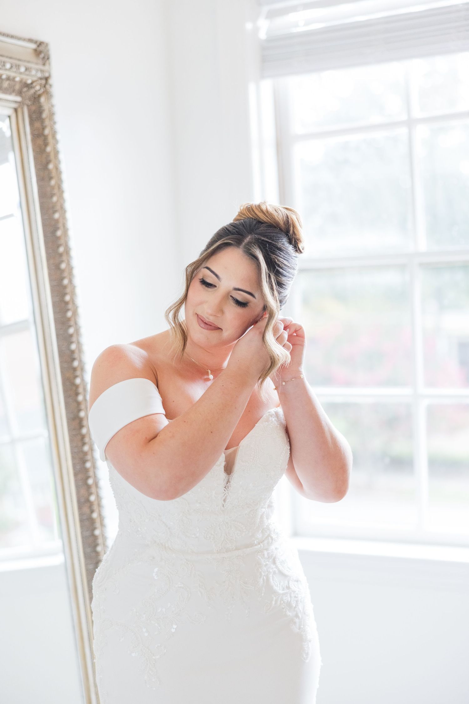A bride in an elegant white gown poses near a decorative mirror in a bright indoor setting.
