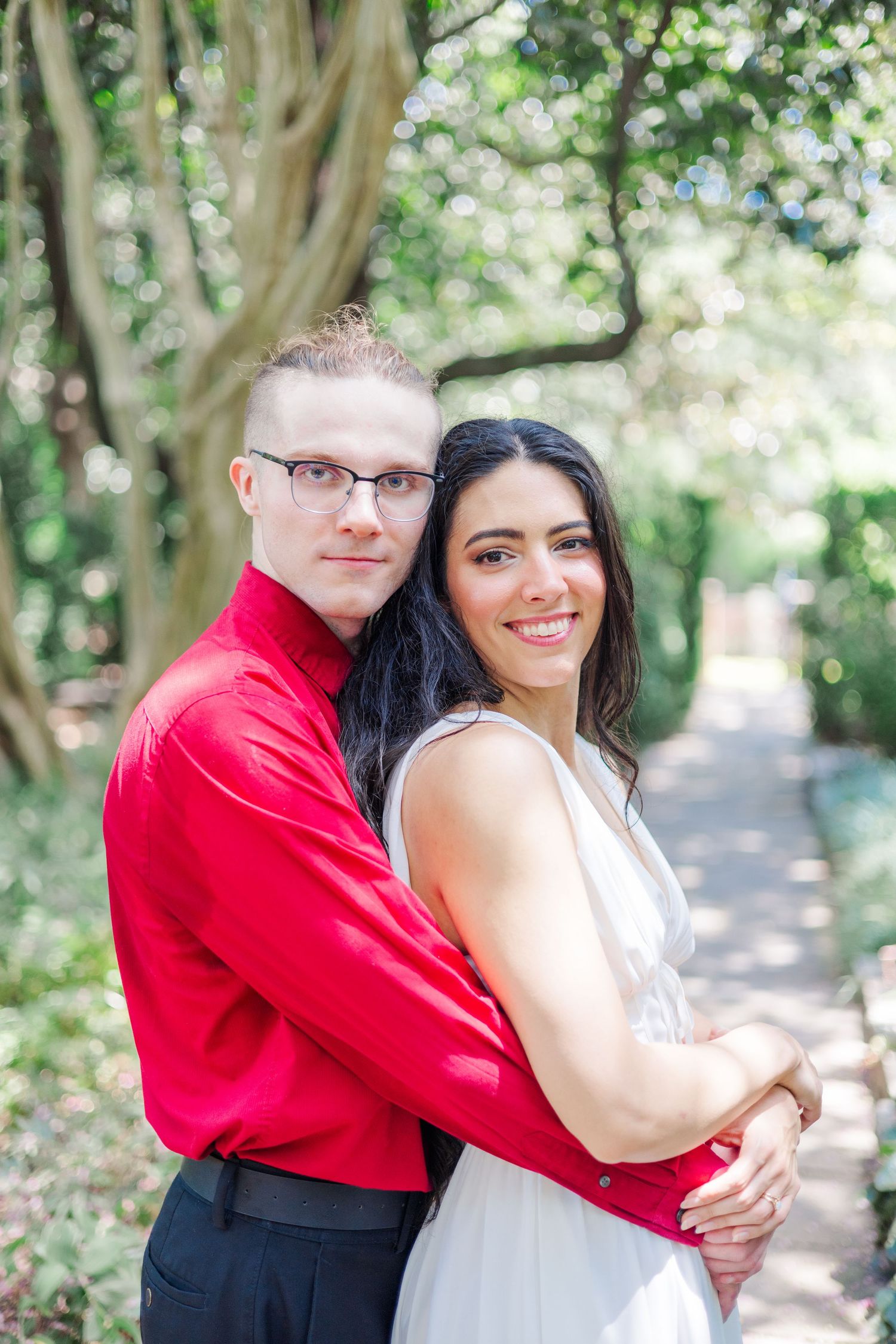 A couple embraces on a tree-lined path, with one wearing a red shirt and the other in a white dress.