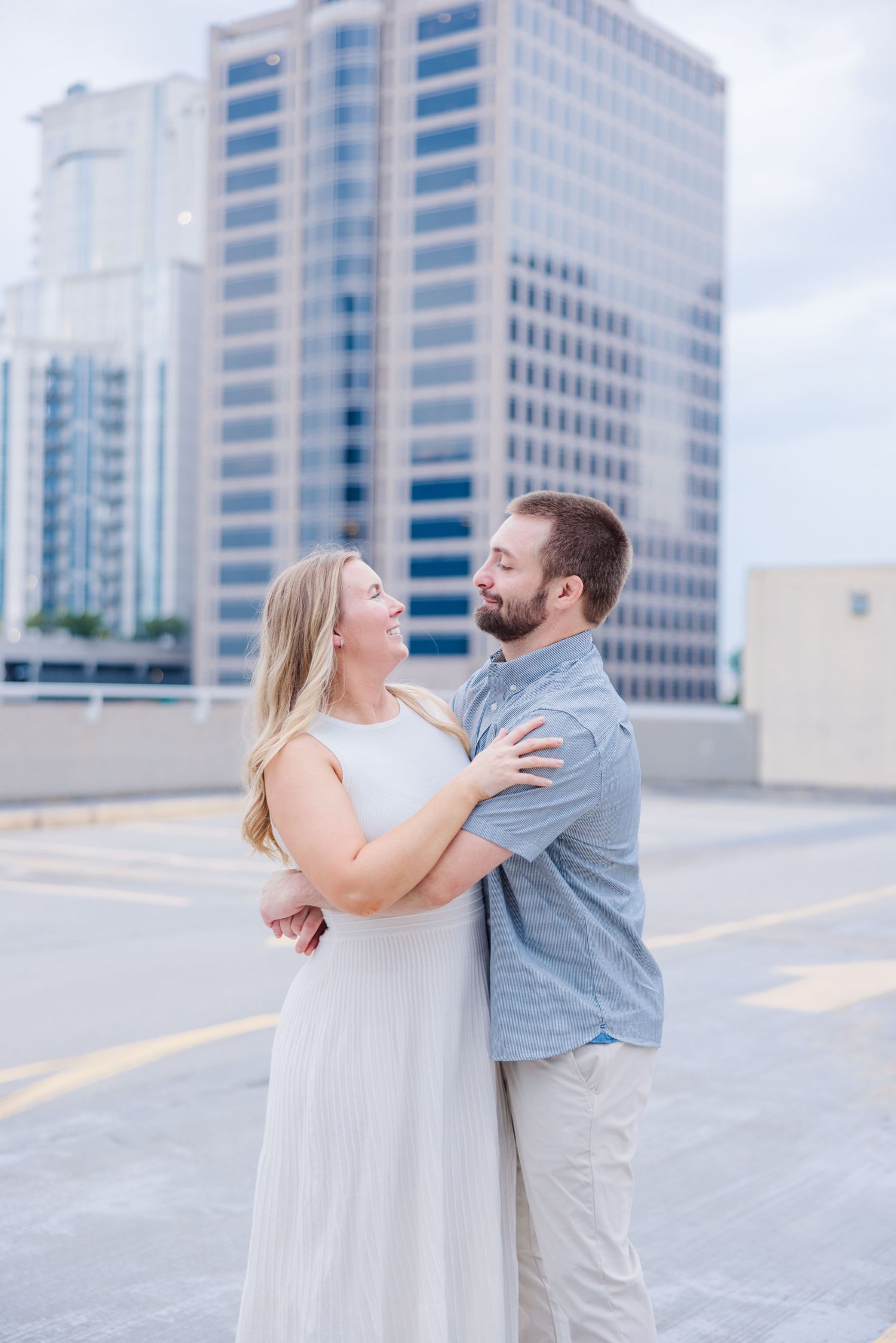 A couple shares a romantic moment on a rooftop parking deck with modern office buildings in the background.