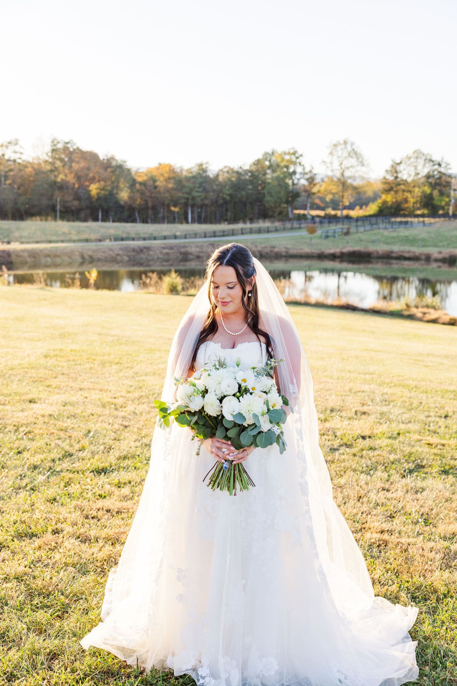A white bridal bouquet with roses and greenery against a sunlit autumn field backdrop.