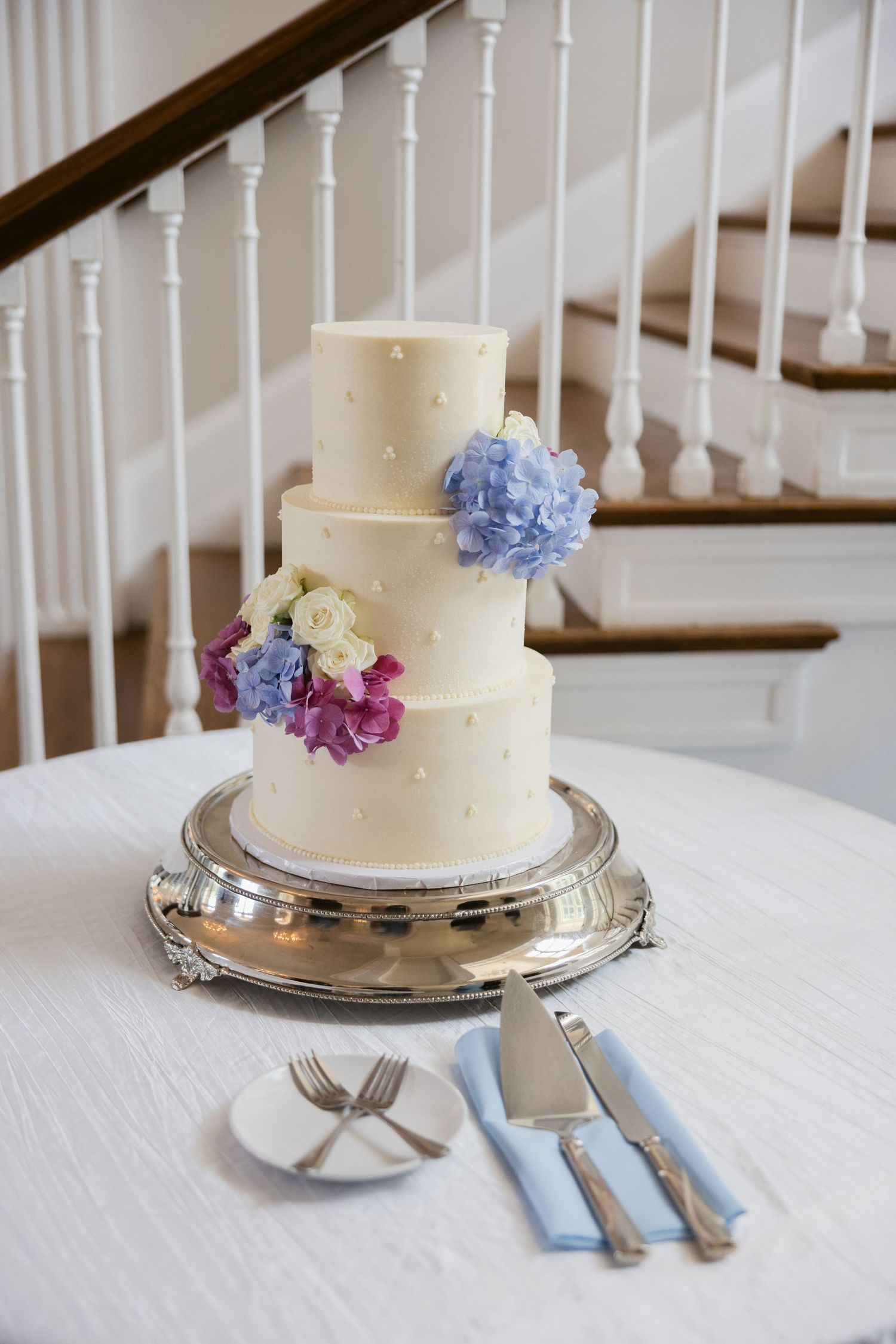 Elegant white wedding cake decorated with delicate blue and purple flowers on a silver platter.