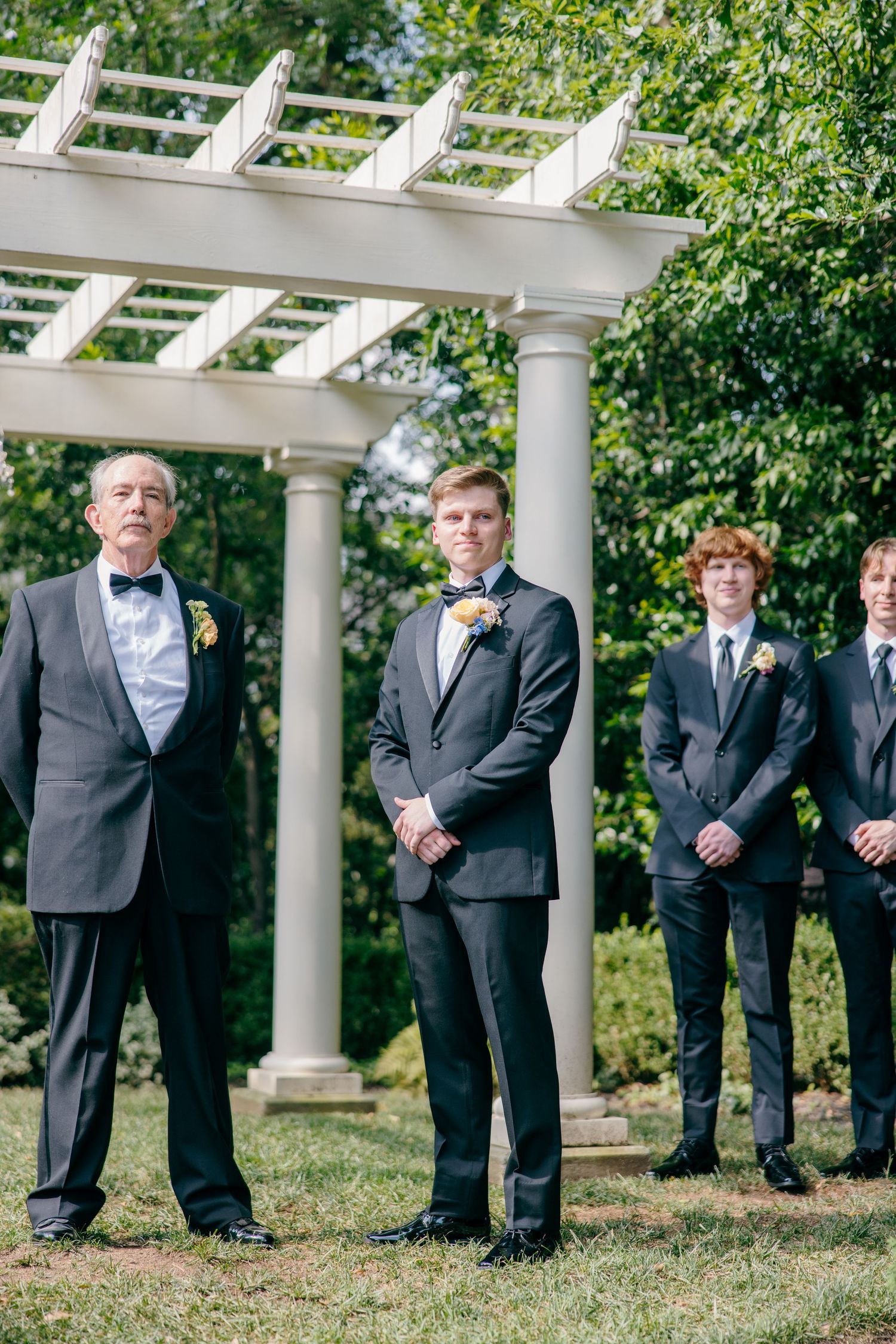 Groomsmen in dark suits stand in formation under a white garden pergola.
