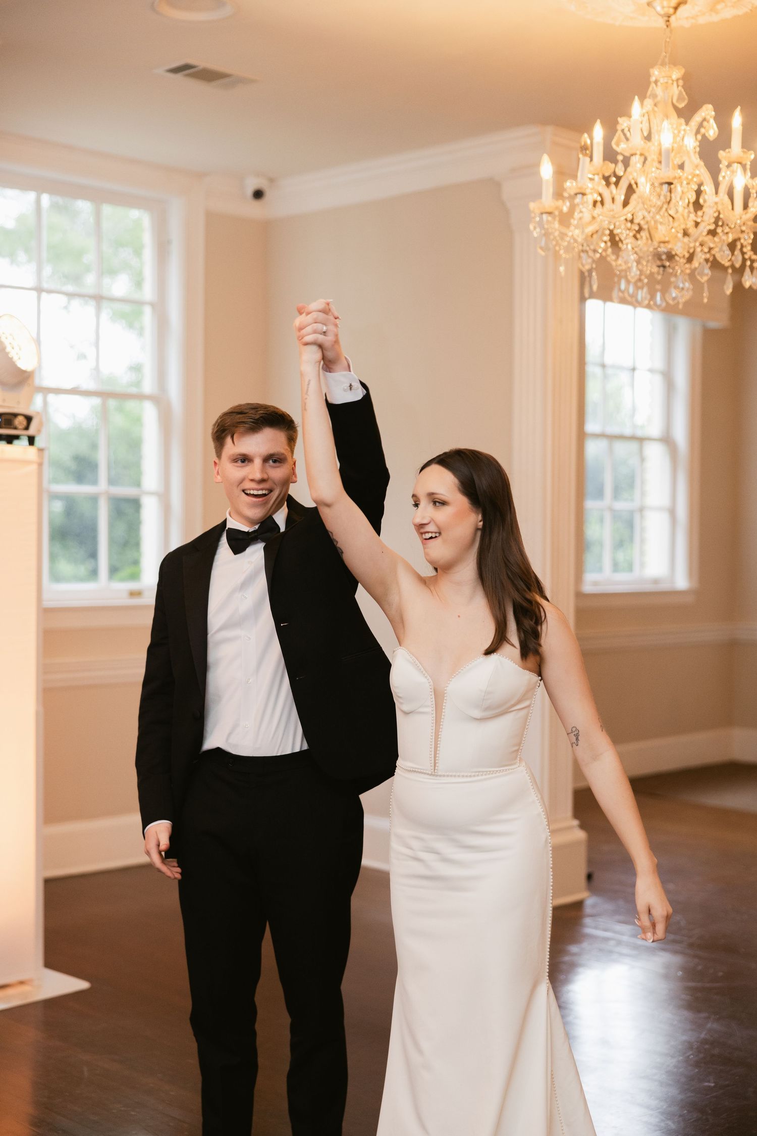 Joyful newlyweds celebrate with raised arms in an elegant room with chandeliers.