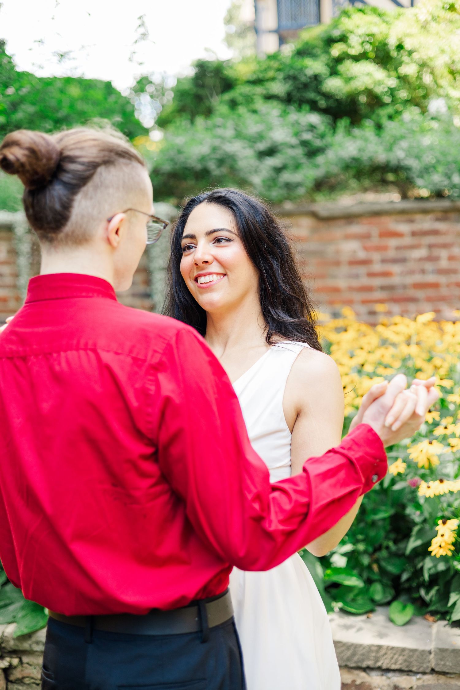 A couple shares a romantic moment in a garden with blooming yellow flowers and a brick wall backdrop.