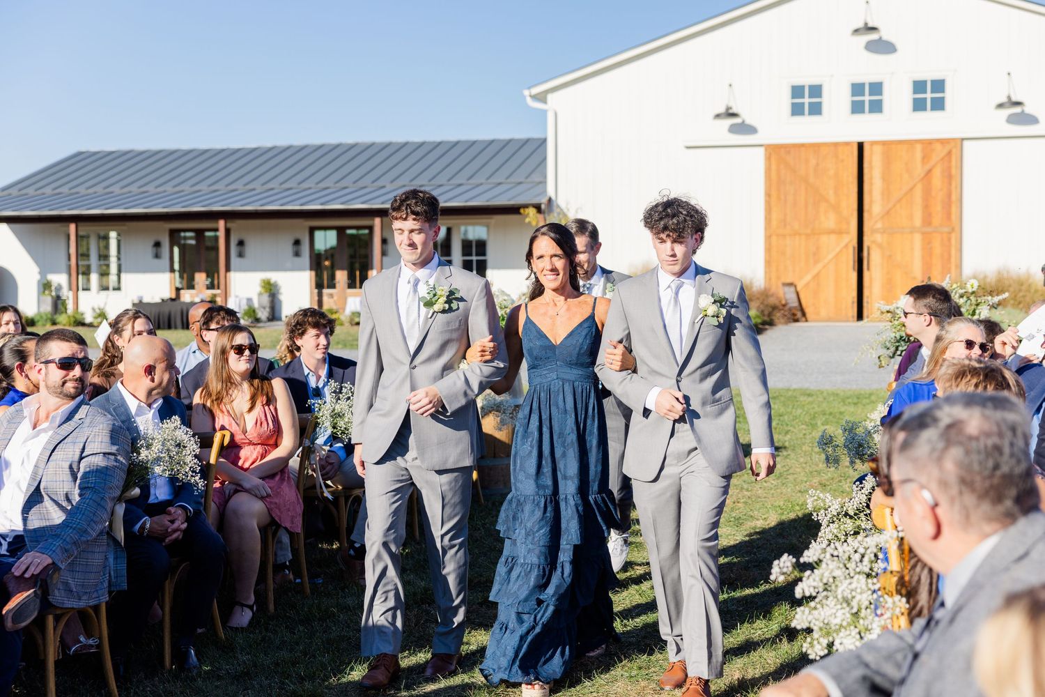 Wedding guests gather outdoors on a sunny day with a rustic building in the background.