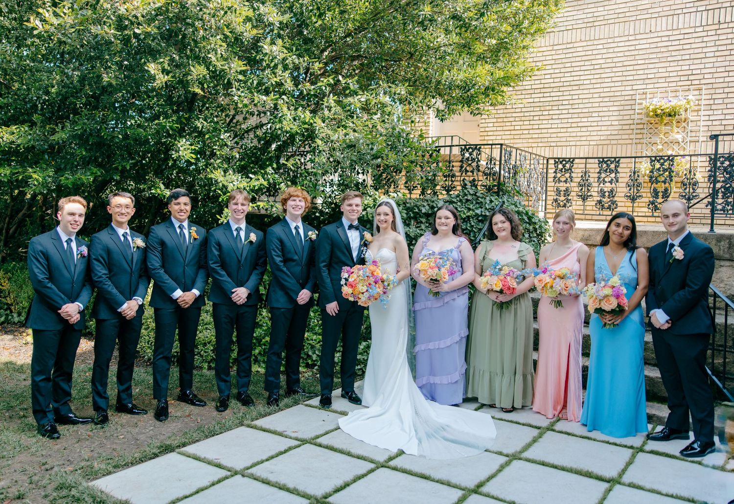 Wedding party lined up outdoors with bridesmaids in pastel dresses and groomsmen in dark suits.