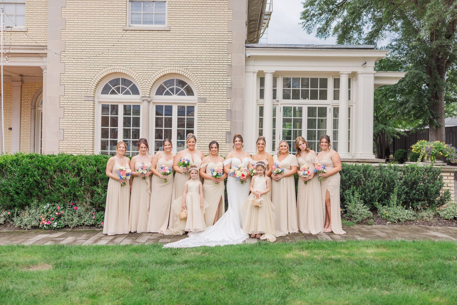 A wedding party in peach and cream colored dresses poses together on a manicured lawn in front of an elegant mansion.