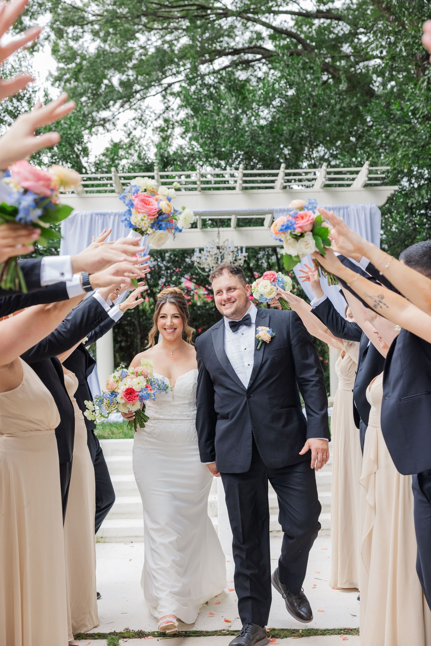 Wedding party celebrating with joyful poses in formal attire under garden pergola.