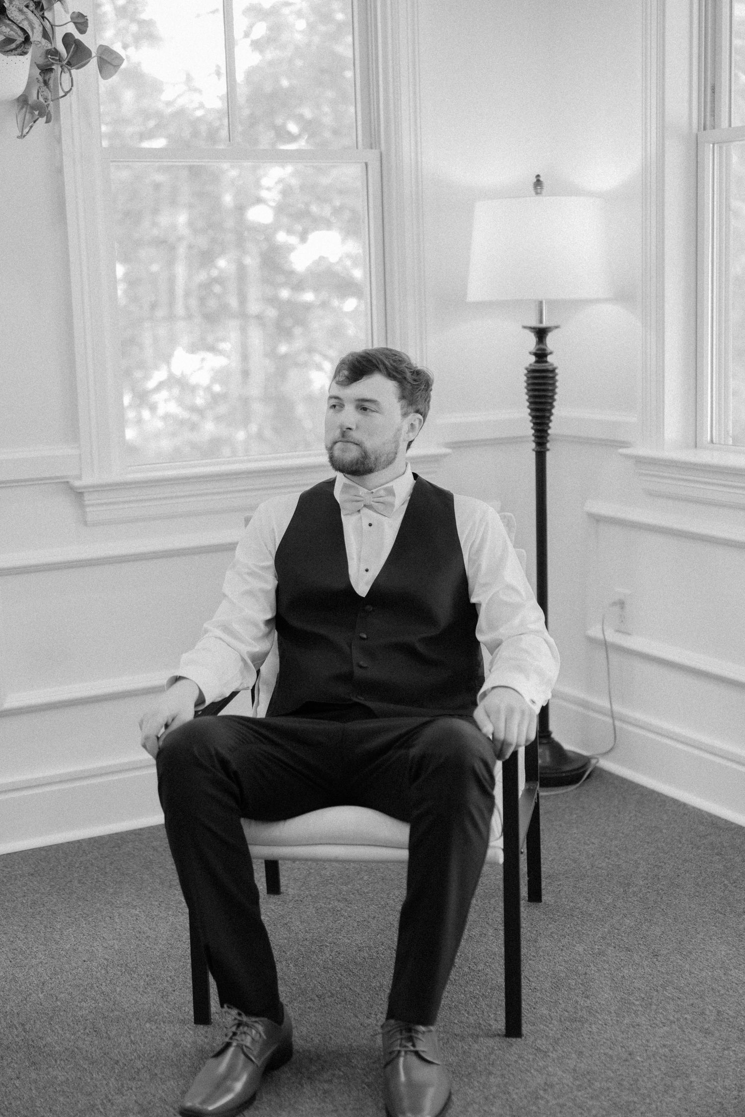 A formal portrait of a groom seated in a chair wearing a vest and bowtie.
