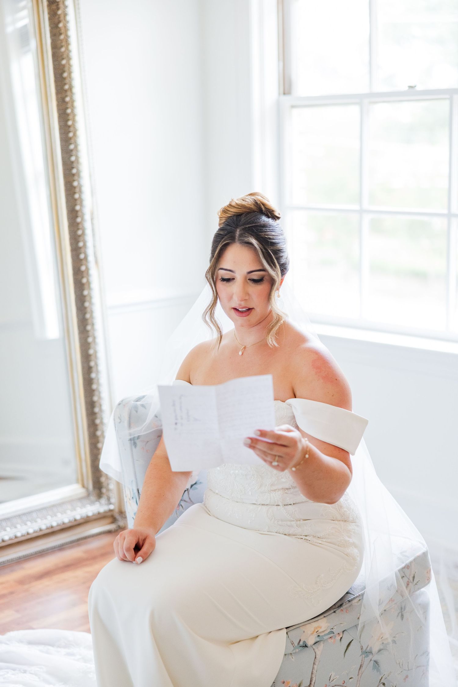 Bride in off-shoulder wedding dress reads letter while seated in bright white room.