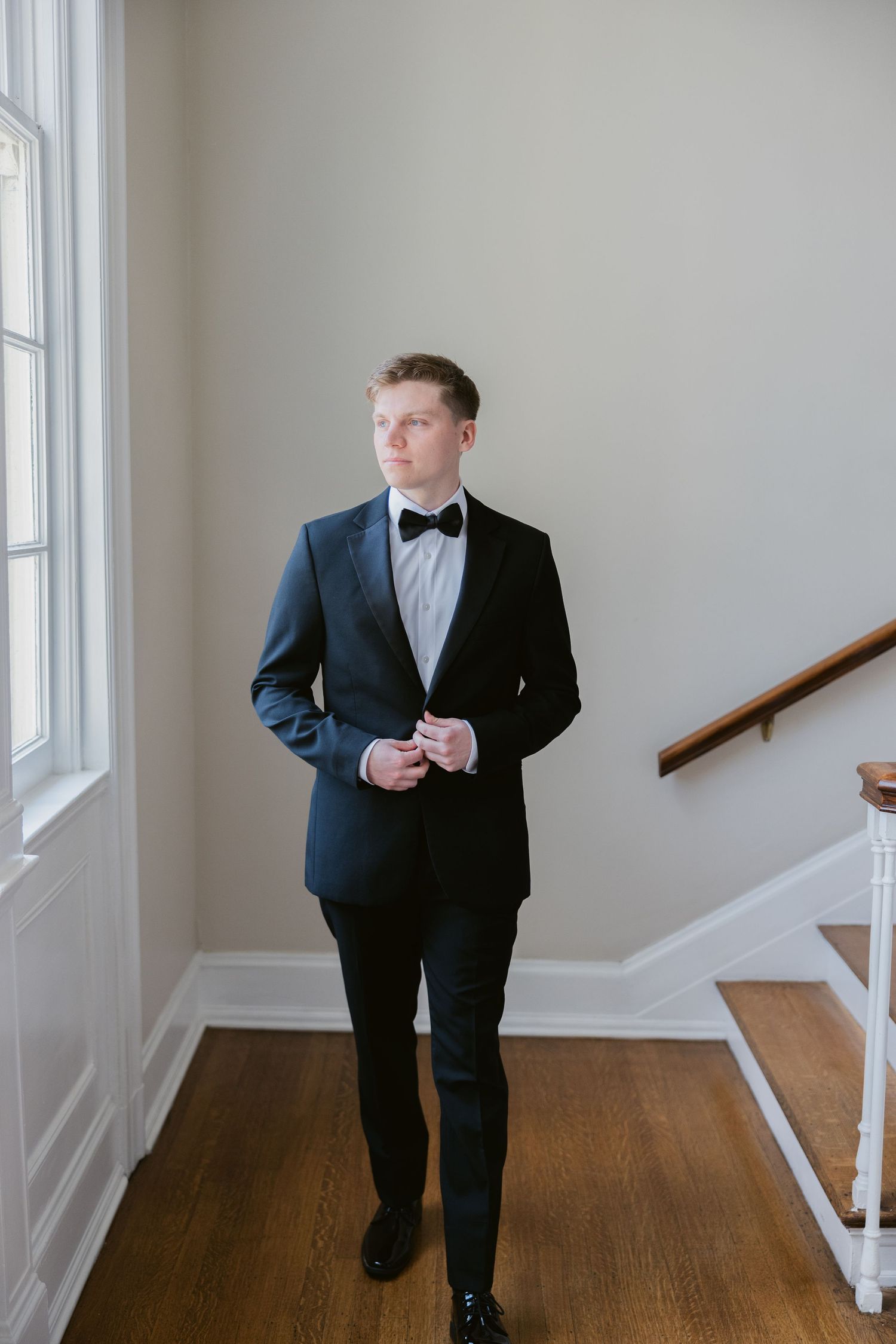 Man in formal black tuxedo standing in an elegant hallway with white walls and hardwood floors.